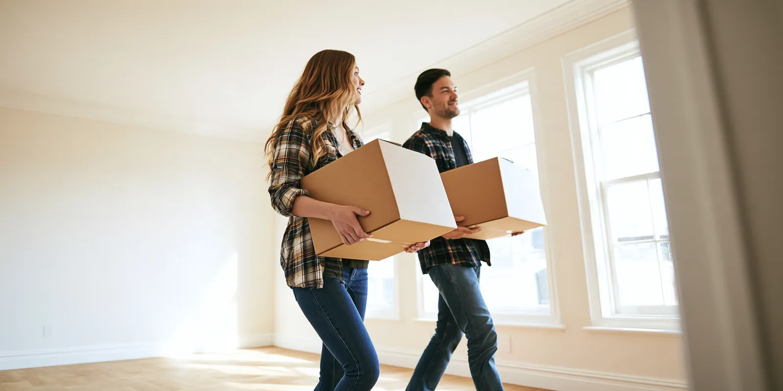Young couple carrying moving boxes into a bright, empty new apartment with big windows, a move paid for partly with an avant lending moving loan