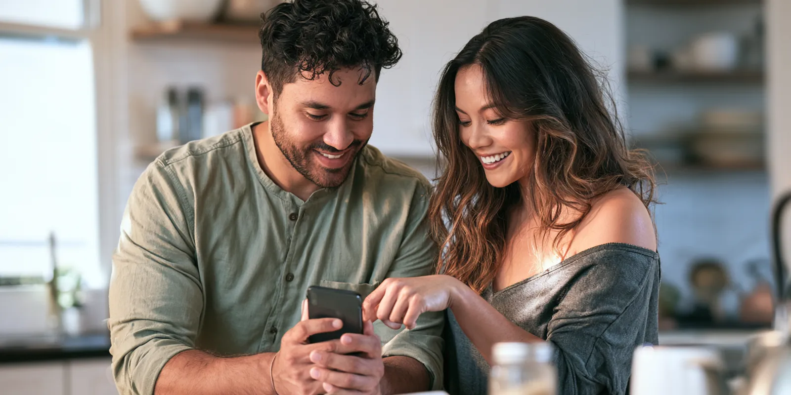 Couple in their 30s smiling with relief as they read an avant lending deposit notification on a smartphone in their kitchen