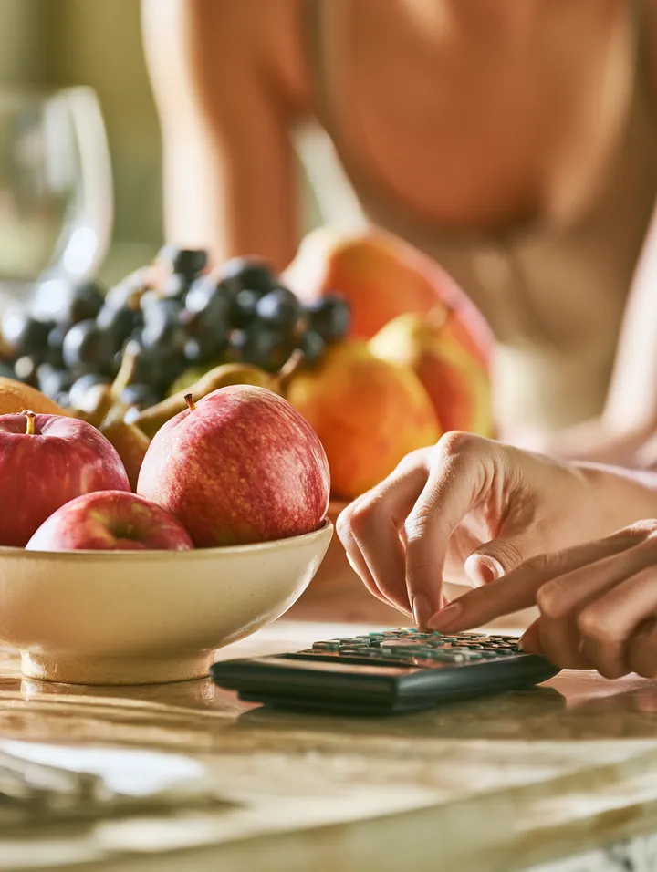 Hands using a slim calculator on a dining table beside a bowl of fresh fruit while comparing card interest with a personal loan