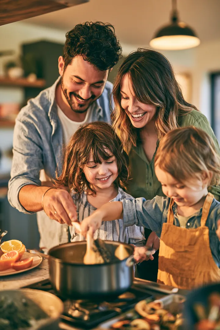 Family of four cooking dinner together in a bright open kitchen, kids stirring a pot, with one card payment instead of three